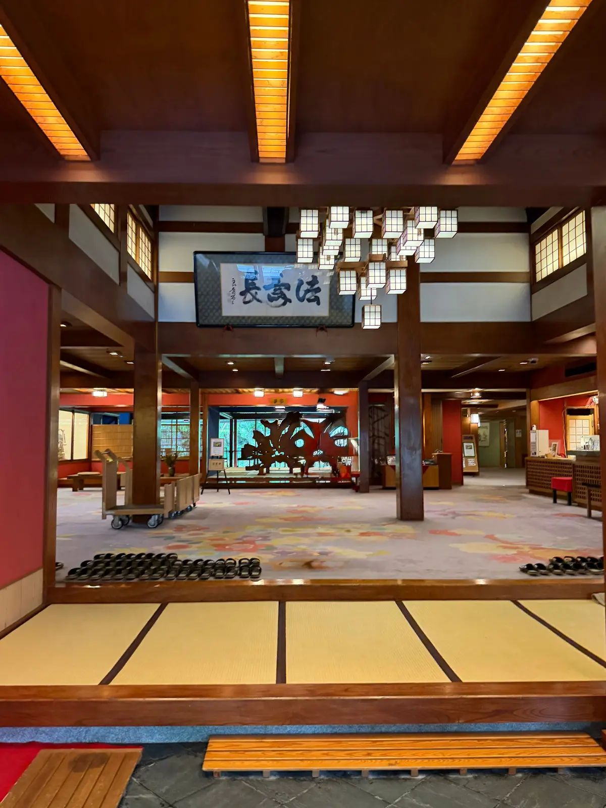伝統的な日本の旅館のロビー。木製の柱や梁、障子、畳敷きの床が特徴的。中央には提灯のような照明器具と、書かれた看板が設置されている。背景には、庭園を望む大きな窓が見える。