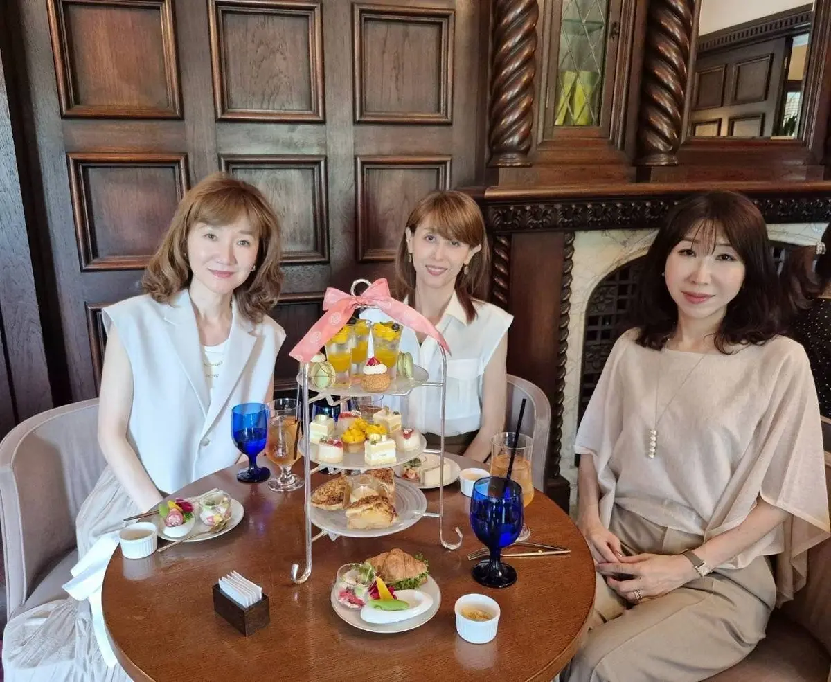 テーブルを囲んで座る3人の女性。中央には三段重ねのケーキスタンドに軽食が並び、青色のグラスが置かれている。背景には木製のパネルと暖炉がある。