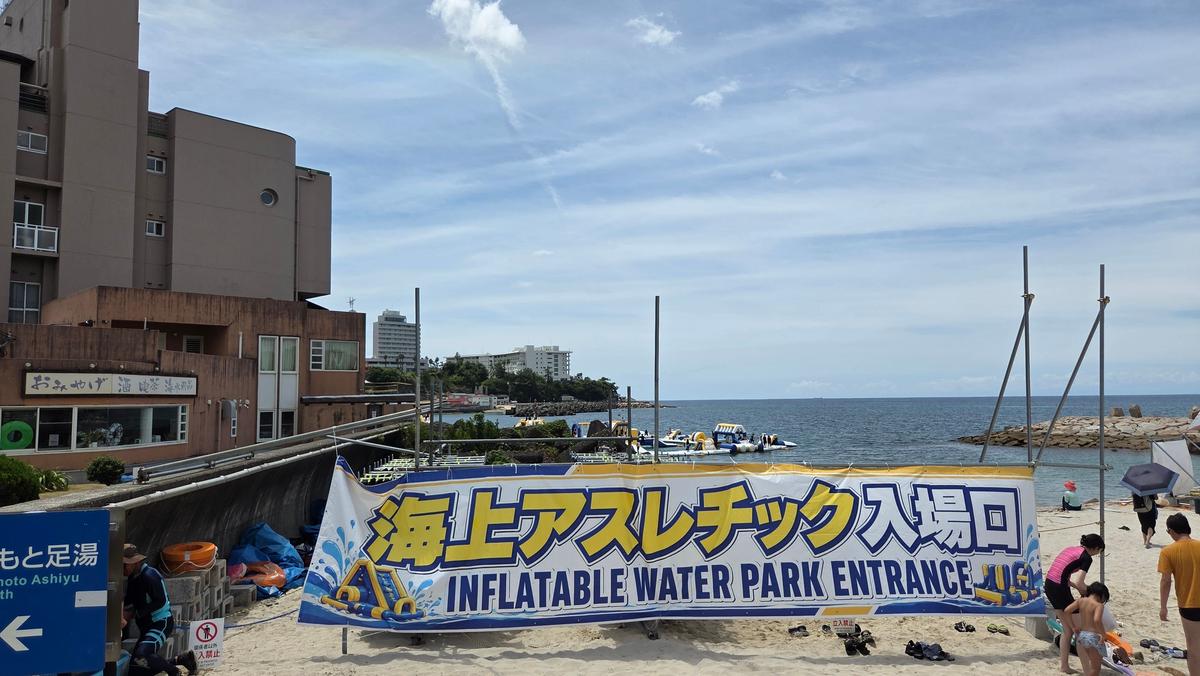 海辺に設置された「INFLATABLE WATER PARK ENTRANCE」と書かれた横長の看板。背景には建物と海、青空が広がり、遠くに波打ち際が見える。