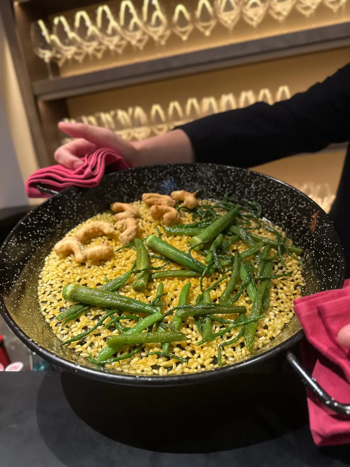 黒いフライパンに、緑豆とエビの炒め物と黄色いライスが盛り付けられています。手はピンクの布でフライパンを持ち上げています。