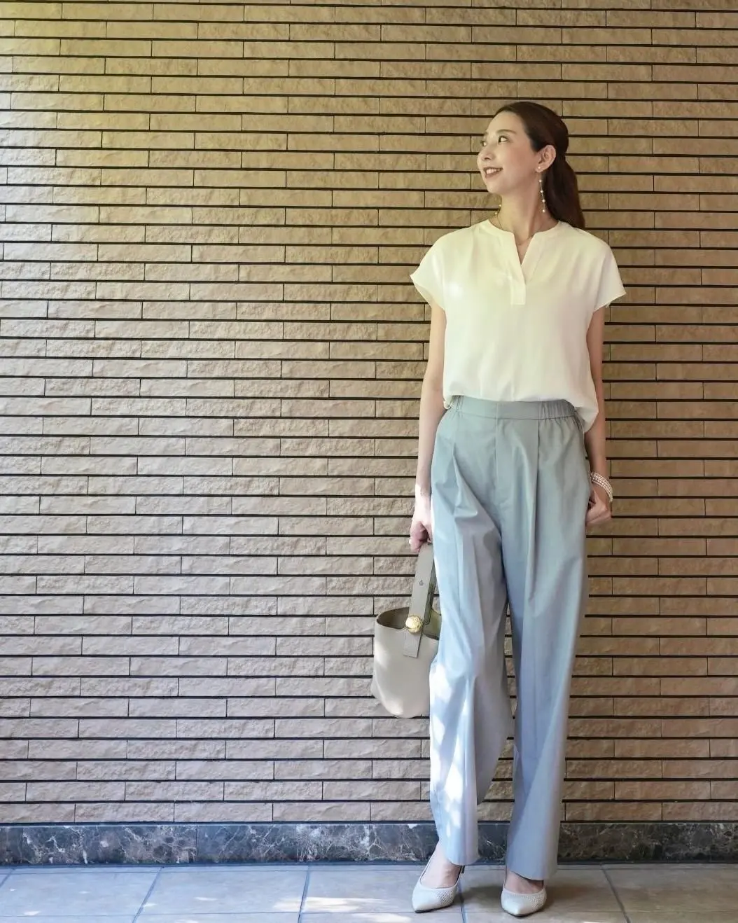 女性がベージュの壁に寄りかかり、軽いグレーのパンツと白の半袖シャツを着て、ベージュのトートバッグを左手に持ち、左を向いて微笑んでいる。