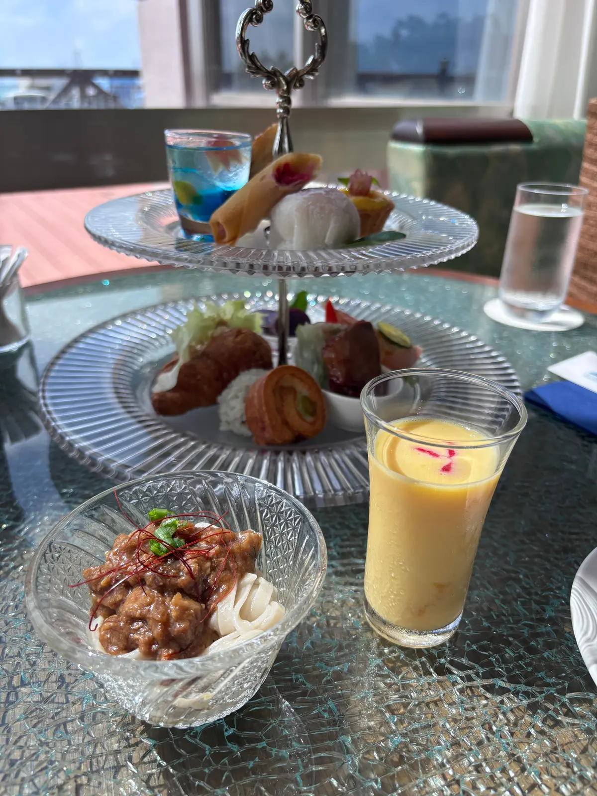 ガラスの2段スタンドに、様々な軽食と飲み物が盛り付けられた様子。最上段にはロール状のパン、球状のスイーツ、青色のドリンク。中段には、肉料理、野菜、リング状の菓子。最下段には、ソースがかかった肉料理とソフトクリーム、黄色いドリンク。