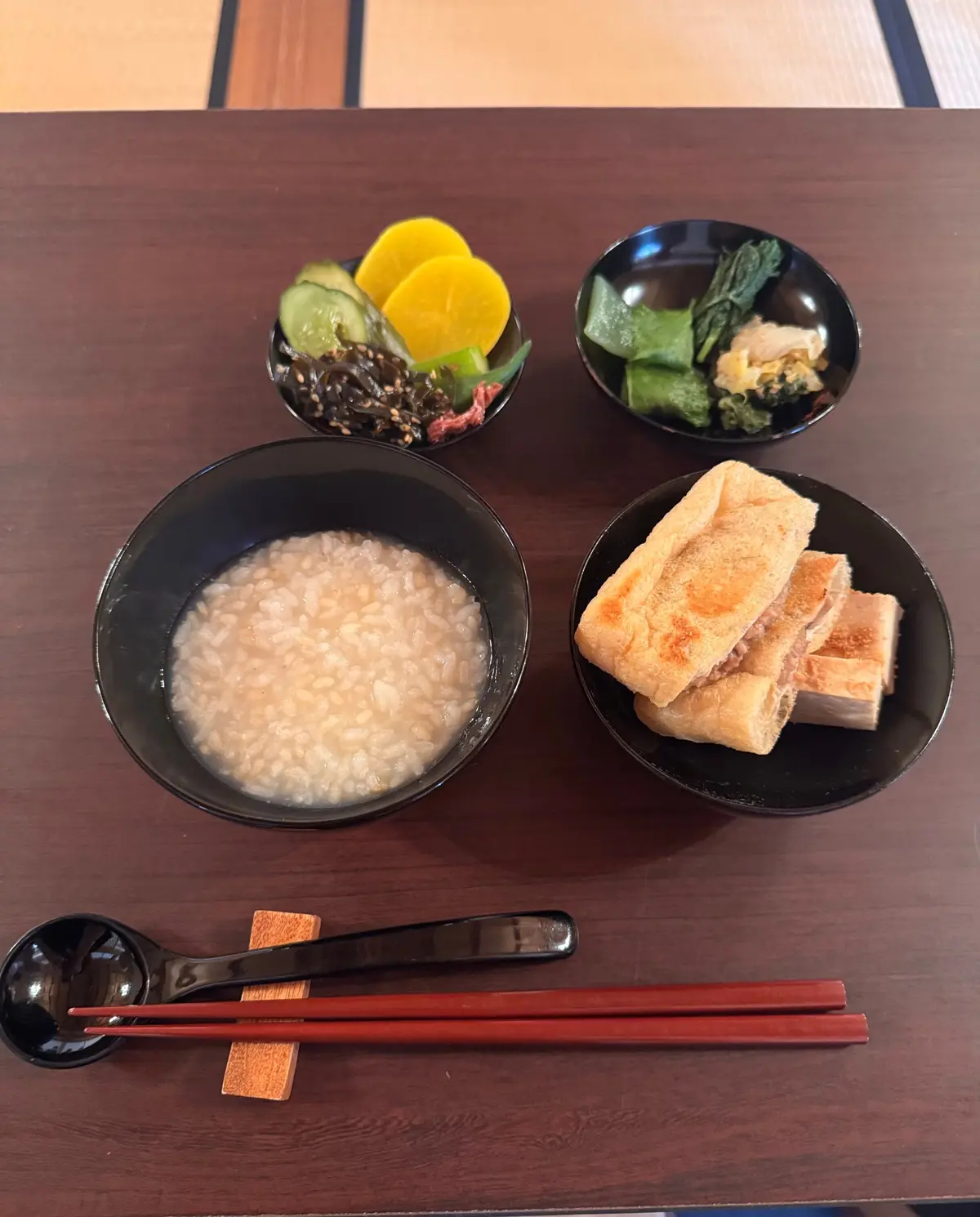木製テーブルに、おかゆ、刺身こんにゃく、野菜の盛り合わせ、大豆肉入り油揚げ、4つの料理が並んでいる。赤い箸とスプーンが置かれている。