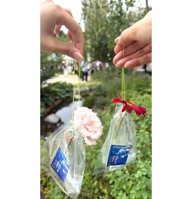 ピンクと赤の kwiaty（クワティ）の花が、透明な袋に入れられ、糸で吊るされている。袋には青いラベルが貼付されており、背景には緑の植物と水辺が見える。