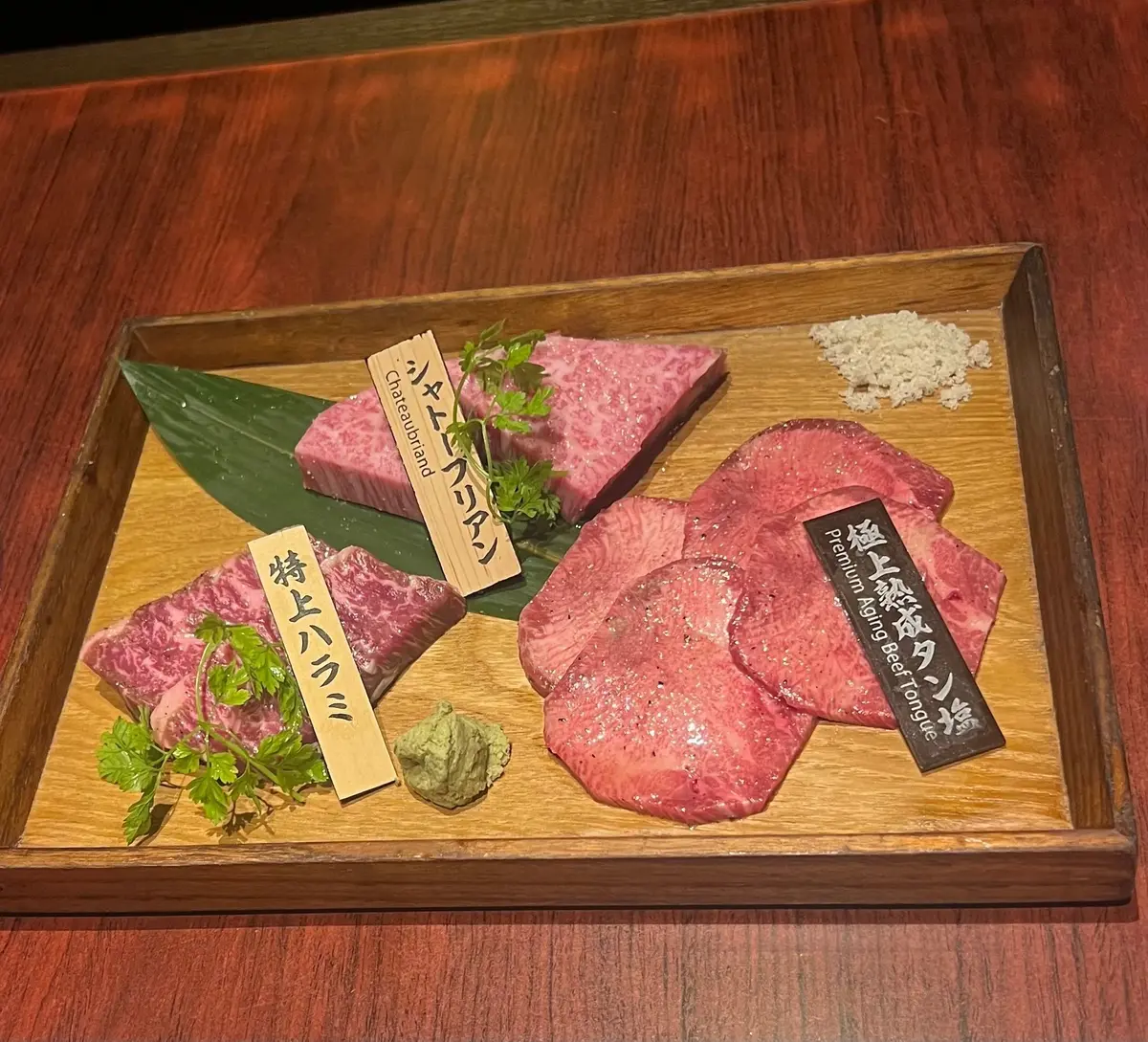 木製のトレーに盛り付けられた、赤身の美しい和牛の肉。シャトーブリアン、牛タン、肩ロースの3種類があり、それぞれ日本語と英語で名前が書かれた札が添えられている。緑の葉と薬味が彩りを添えている。