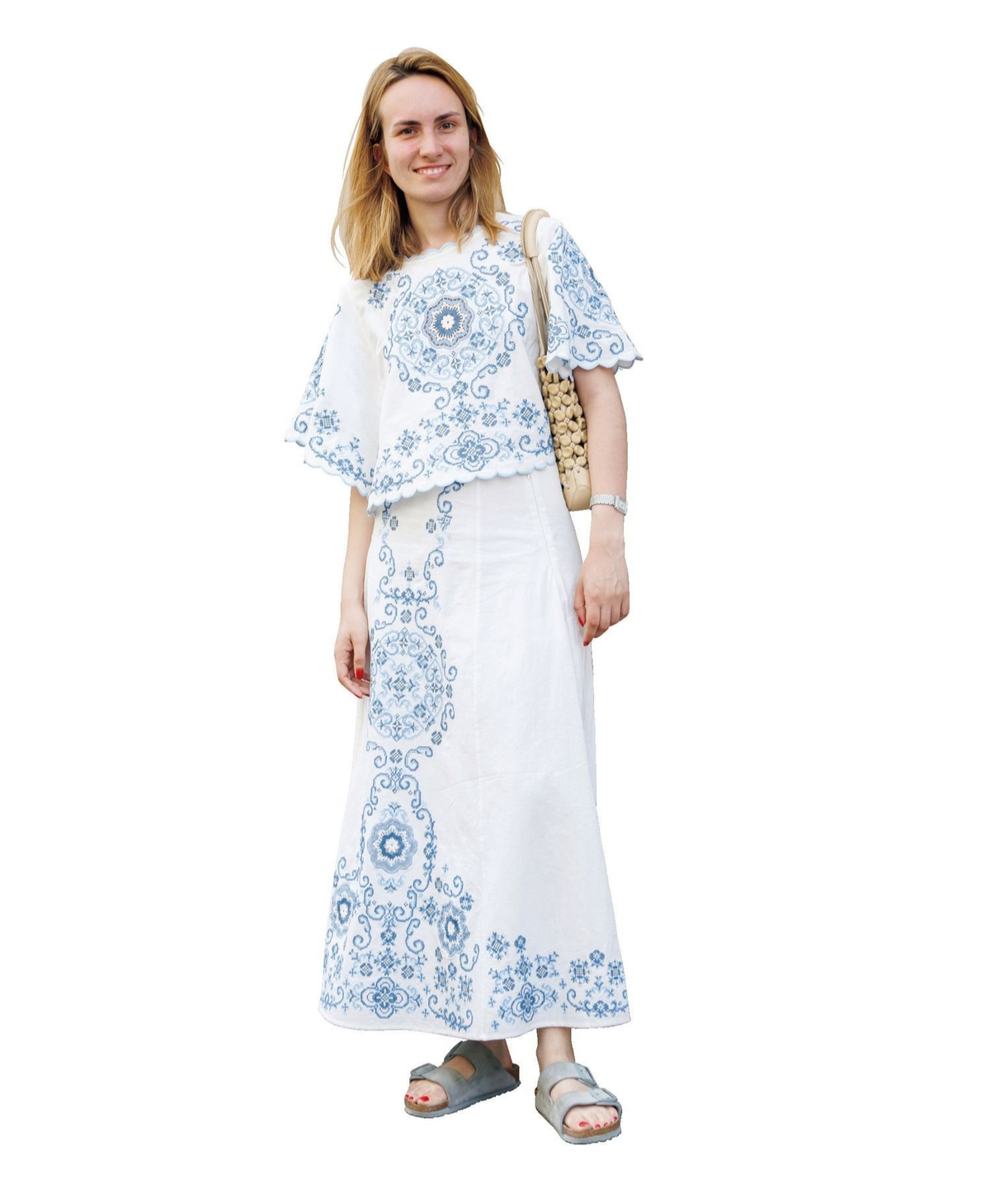 白い刺繍のトップスとロングスカートを身につけた女性。淡い青色の花柄刺繍が施され、右手に麦わらのバッグ、足元にはサンダル。笑顔でこちらを見ている。