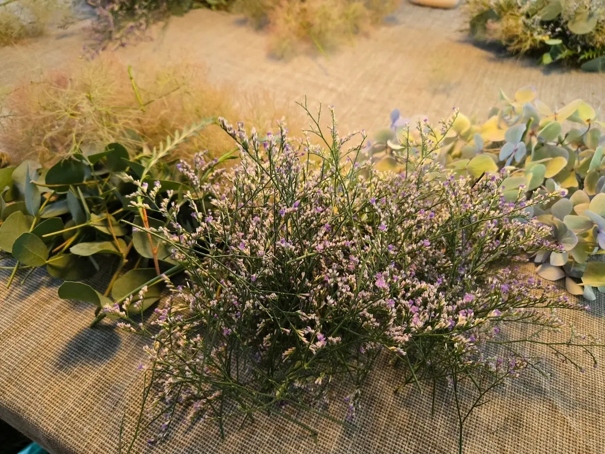 麻布の上に、紫色の小さな花が咲いた植物と、緑のユーカリの葉、淡い色合いの花束が置かれている。