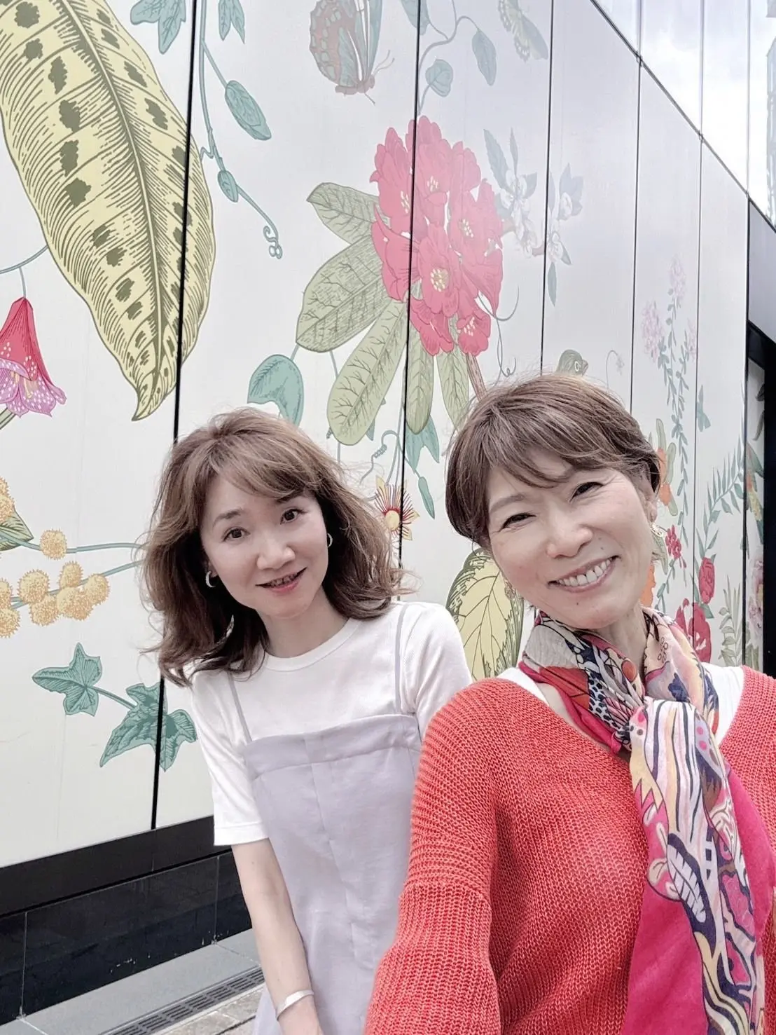 花の模様が描かれた壁の前で、若い女性と年配の女性が並んで笑っている。