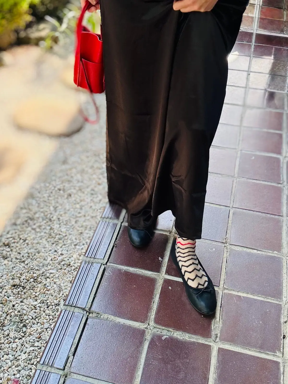 黒のズボンに黒のフラットシューズ、赤と黒の縞模様の靴下を履いた人の足元。赤のハンドバッグが部分的に見える。