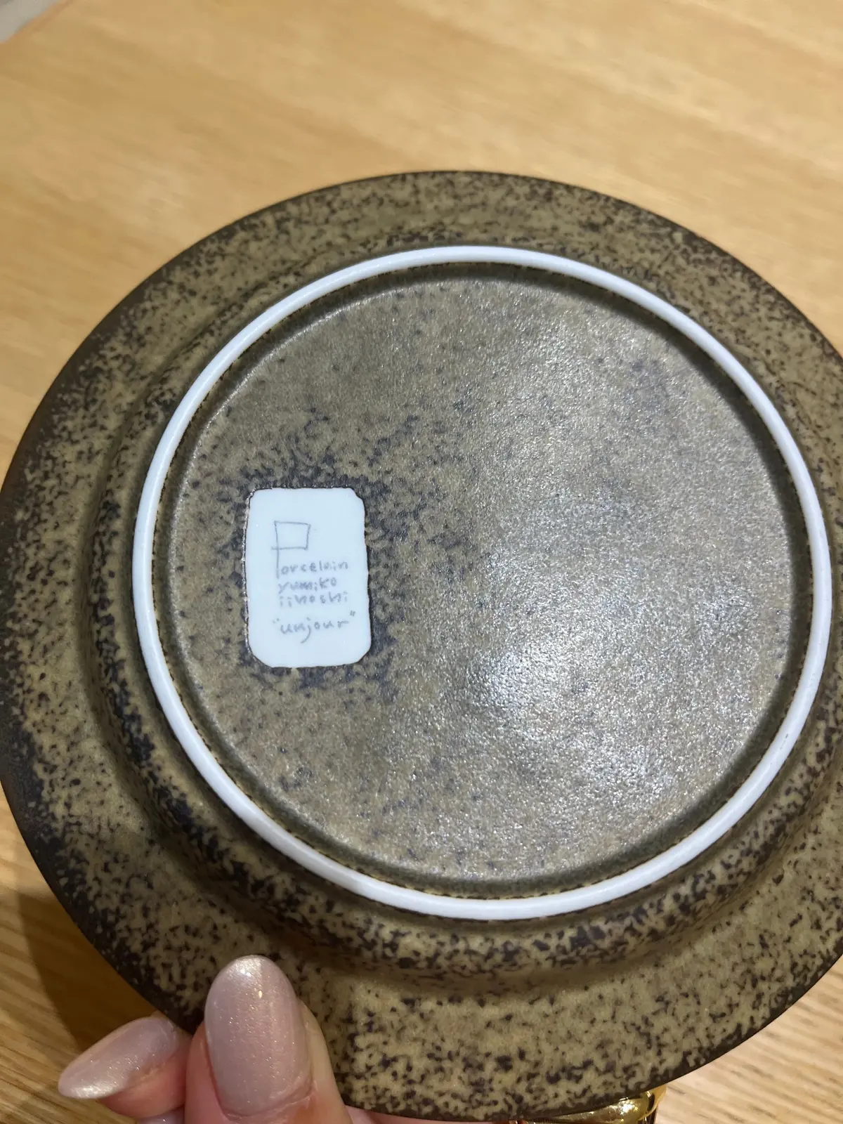 木製のテーブルに置かれた陶器の皿の裏面。表面は茶色と黒のまだら模様。中央には白いラベルがあり、「porcelain yumika inoshita 'unjour'」と手書きで記載されている。縁は白く、皿の形状は円形。