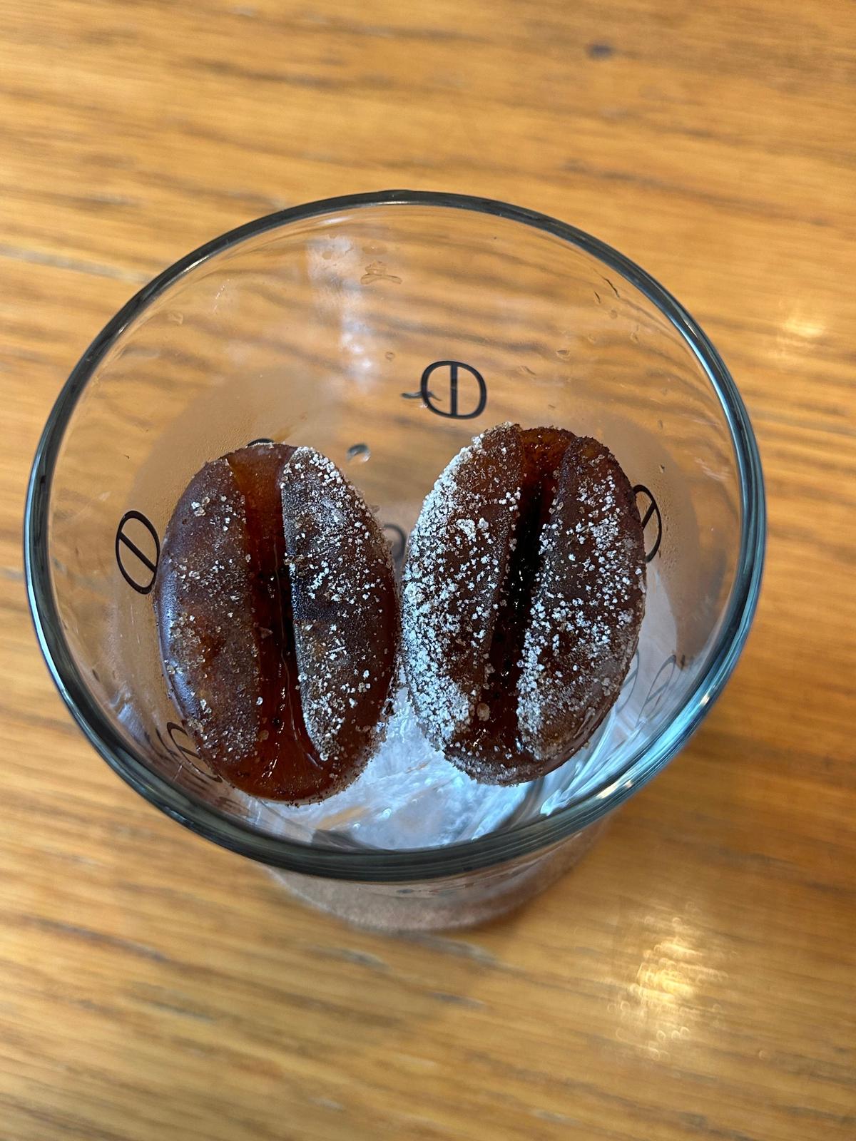 透明なグラスに氷水が浮かび、その中に3つのチョコレートドーナツが並んでいる。ドーナツは楕円形で、表面に白い粉砂糖がふられている。