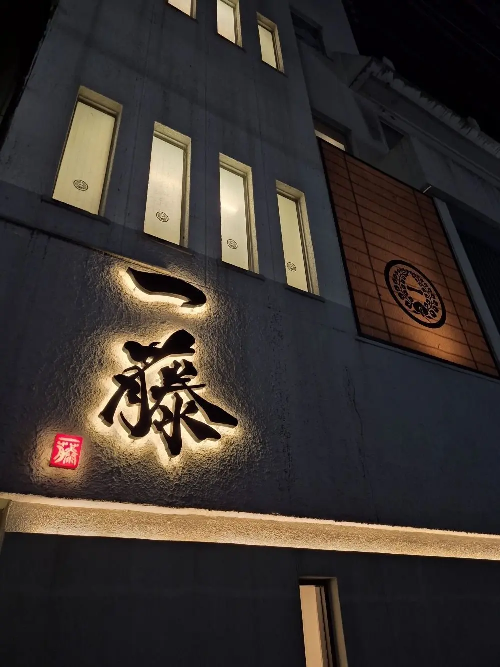 夜の建物に設置された、黒い文字で「藤」と書かれた看板。赤い小さなマークが看板の下に配置されている。窓が複数あり、光が差し込んでいる。