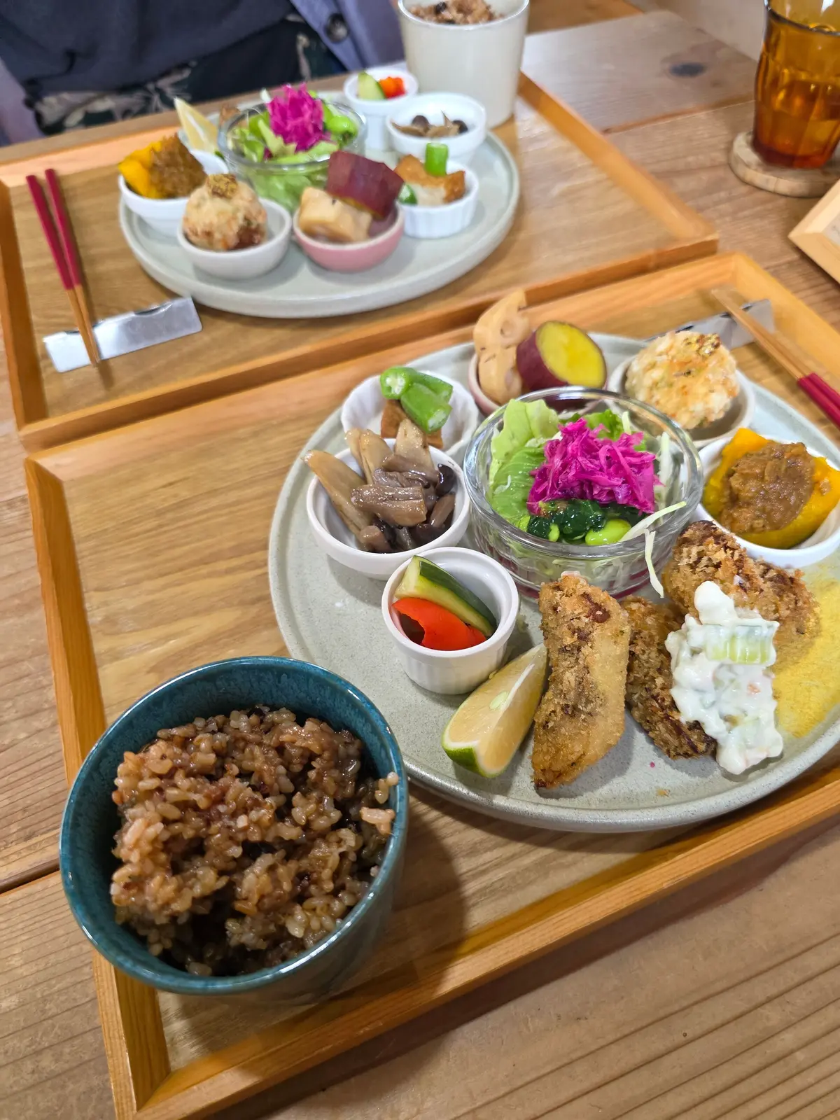 木製のトレイに盛り付けられた、様々な小鉢料理。玄米ご飯、きのこ、サラダ、魚のフライ、和え物など、彩り豊かで多様な食材が並んでいる。背景には似たような盛り付けの料理が置かれている。