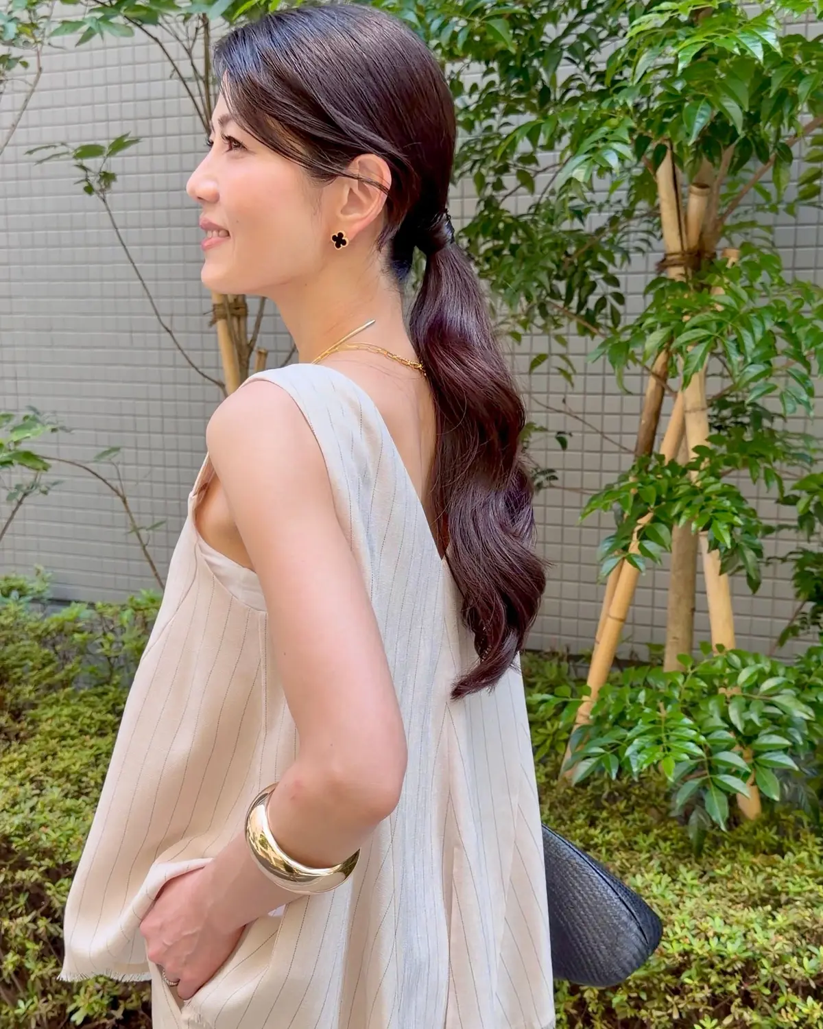ベージュのストライプのタンクトップを着た女性が、横を向いてこちらを見ている。茶色の髪は後ろでポニーテールにまとめられ、ゴールドのアクセサリーを身に着けている。背景には緑の植物と白い壁がある。