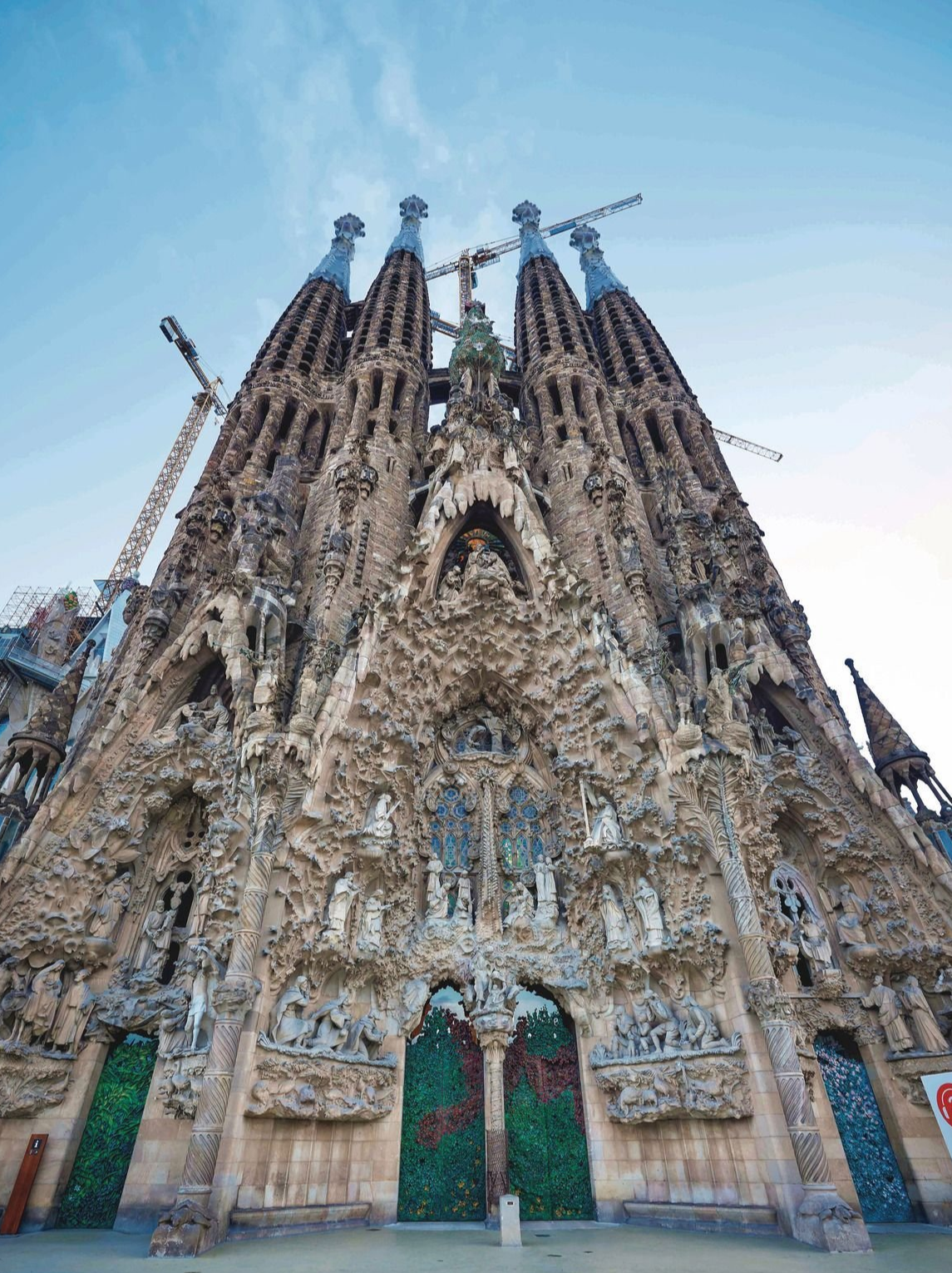 スペイン、バルセロナのサグラダファミリア正面。高さ約50mの尖塔が8つ立ち並び、石造りの複雑な彫刻が施されたファサードは、自然の有機的な形を思わせる。建設中のクレーンが上空に設置されている。