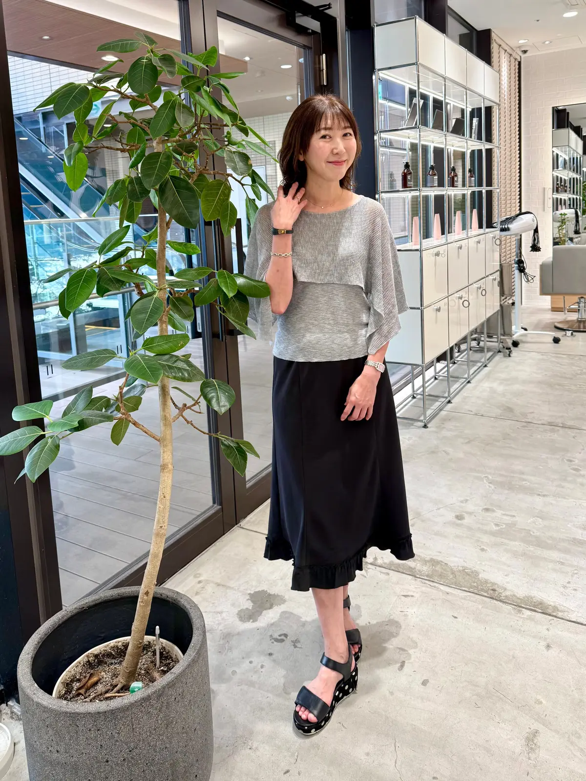 グレーのニットトップスに黒のフレアスカート、黒いサンダルを着用した女性が立っている。左手に手を添え、右手をスカートに当てて微笑んでいる。背景には窓と、白い棚や椅子が見える。足元には植木鉢に入った植物がある。