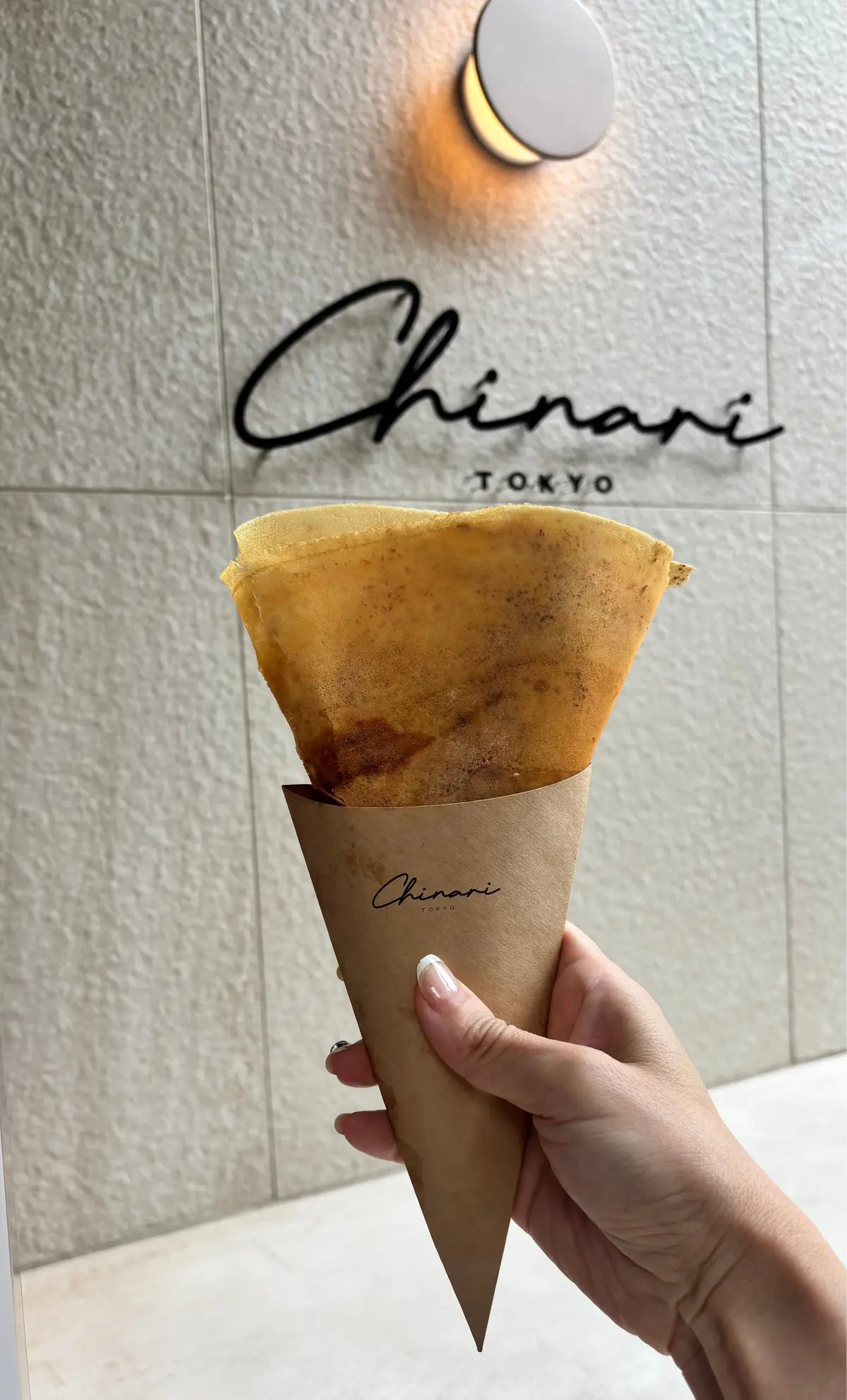 Chinarari Tokyoのロゴが入った茶色のコーンに、焼き立てのクレープが挟まれている。手で持ち、背景にはグレーのタイル壁がある。
