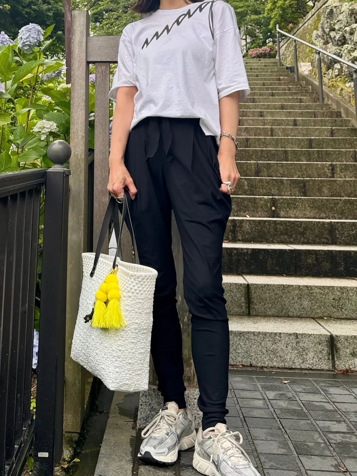 白のTシャツと黒のズボンを着た女性が、階段の手すりにつかまりながら、白い編みかごバッグを左手で持ち、黄色いタッセルが付いています。背景には緑の植物と紫の花が咲いています。
