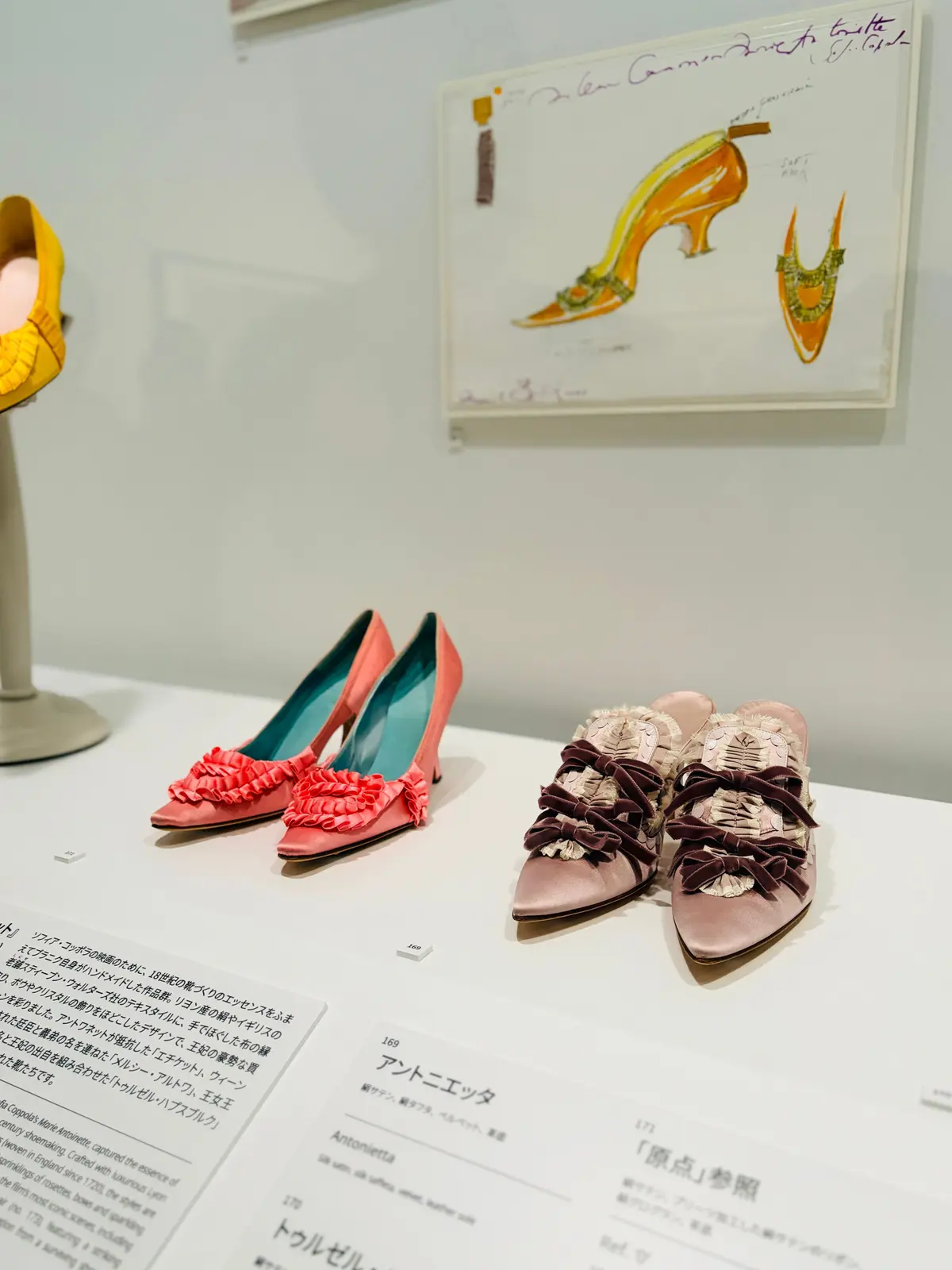展示ケース内の靴の展示。黄色、ピンク、紫、茶色の装飾が施された靴が並び、そのデザインスケッチが壁に貼られている。背景には日本語のテキストが見える。