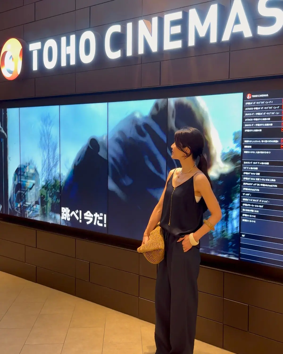 映画館TOHO CINEMASの入り口で、若い女性が映画のポスターを眺めている。ポスターには白と黒の縞模様の動物（おそらくシマウマ）が写っている。女性はベージュの帽子をかぶり、黄色の服を着ている。