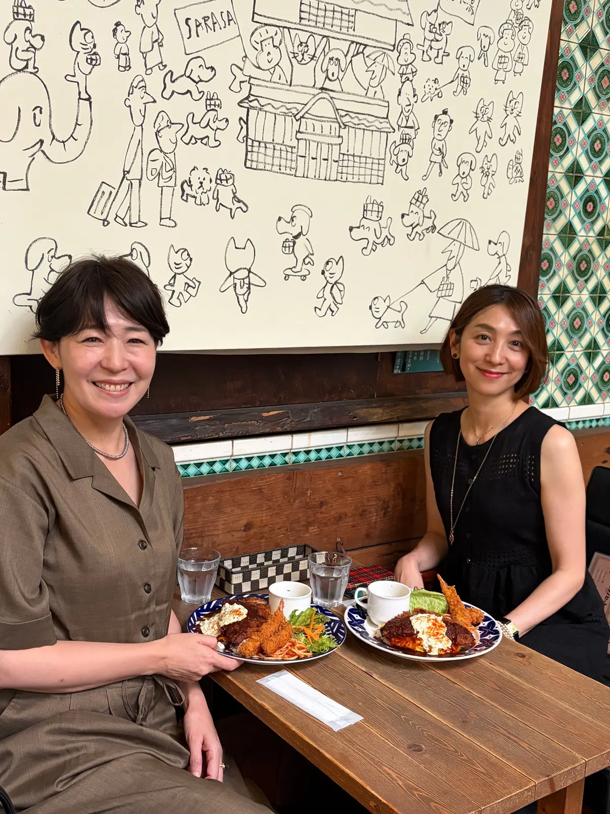 二人の女性が木のテーブルに座り、フライドポテトとサラダの料理を前にして笑っている。背景には犬や人々の漫画が描かれた壁がある。