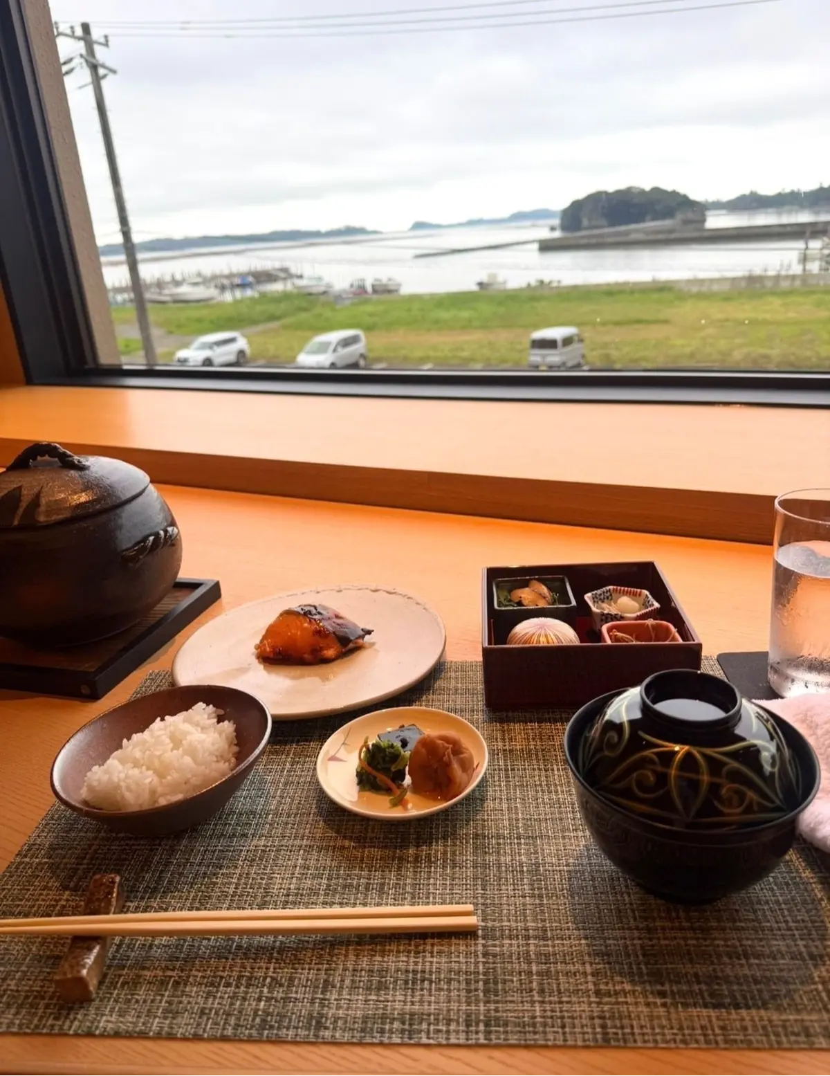 窓から海を見渡せる席で、和食の定食が並んでいる。ご飯、焼き魚、お味噌汁、小鉢、お漬物が盛り付けられている。木製のテーブルに敷かれたグレーのマットが目立つ。