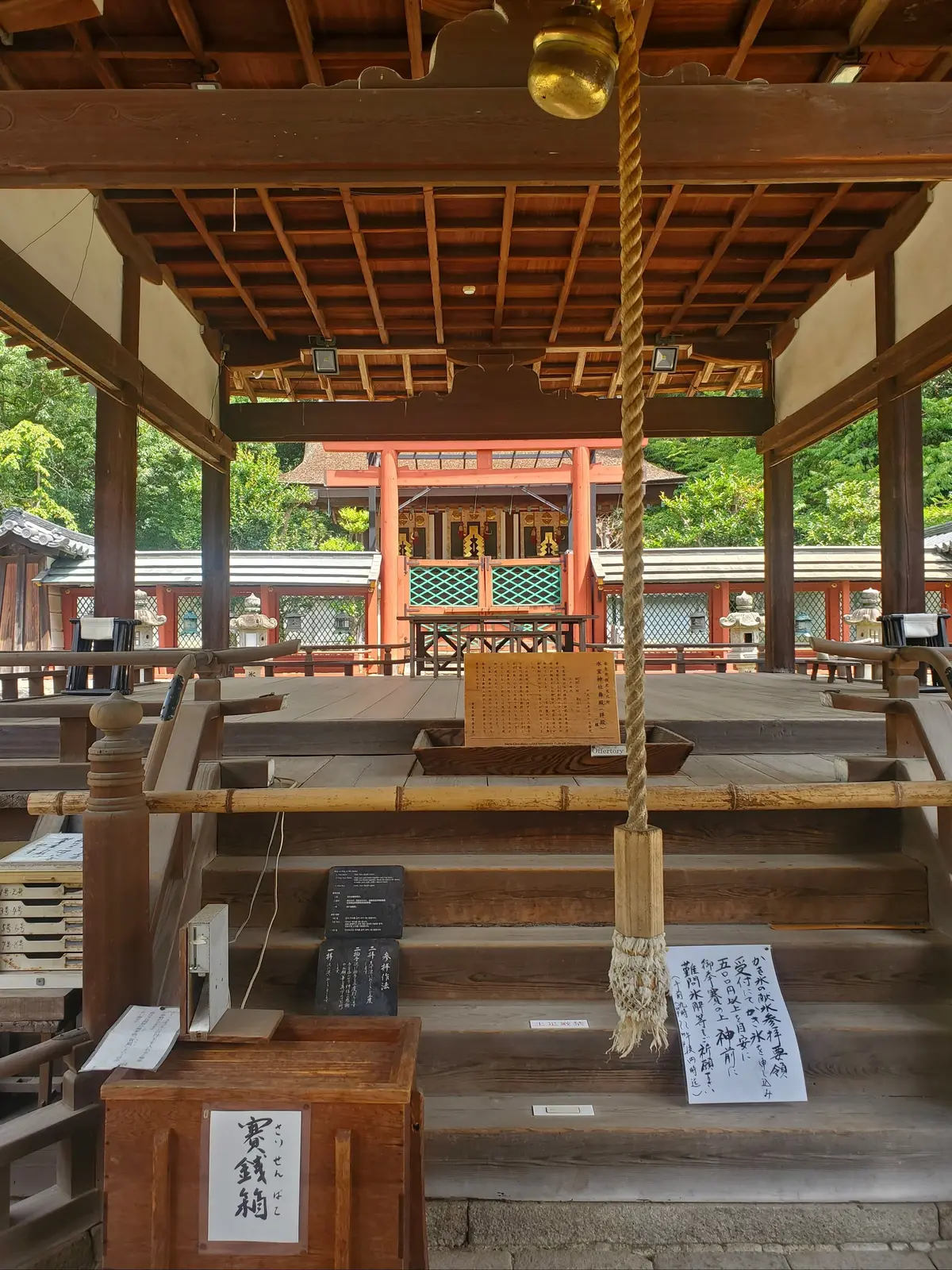 木造の屋根の下、中央には大きな鈴が吊るされている。階段を上がると、朱色の社殿が見える。周囲には緑豊かな木々が茂り、伝統的な日本の風景が広がる。