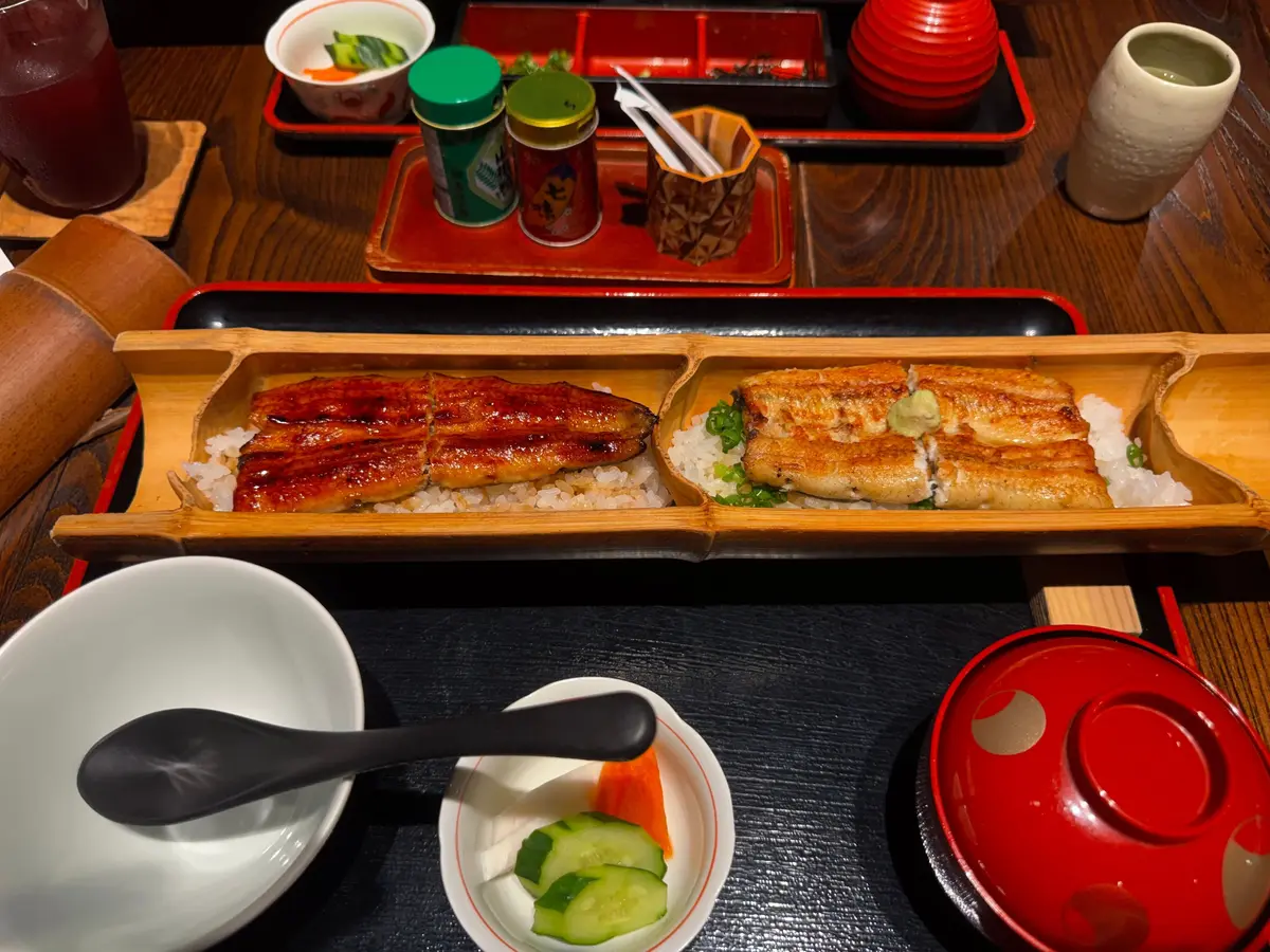 長方形の木製お盆に、鰻重が乗っている様子。左側は鰻が照り焼きされており、右側は蒲焼き風。ご飯と刻み海苔が添えられている。調味料と、お新香、汁物も写っている。