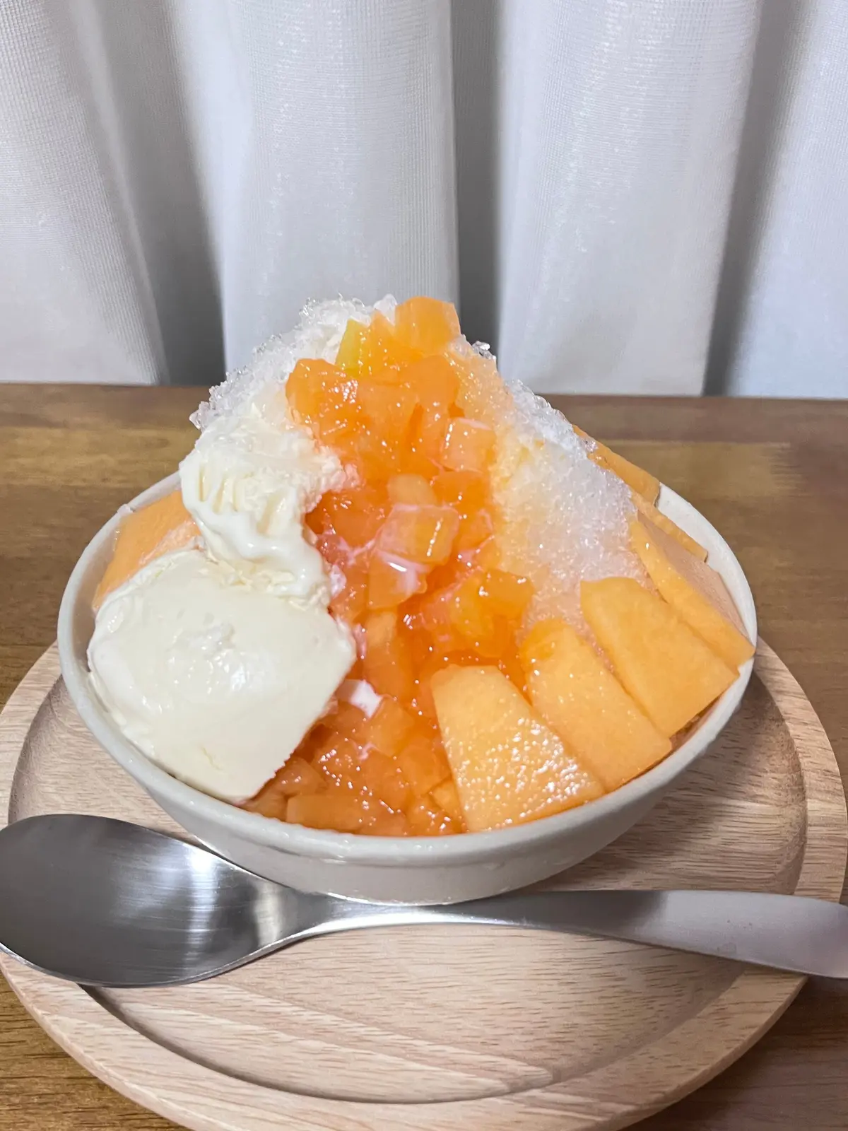 白くて丸い器に盛り付けられた、かき氷とメロン、アイスクリームのデザート。かき氷の上に、オレンジ色のメロンが散らばり、左には白いアイスクリームが添えられている。木製のトレーに置かれ、スプーンが添えられている。