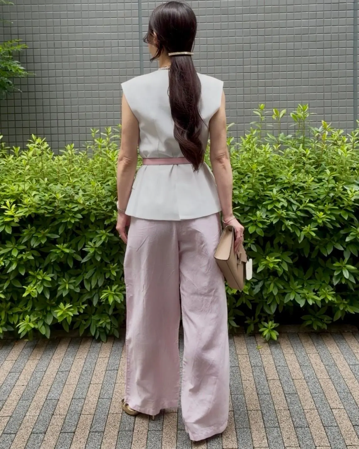 白のノースリーブトップとピンクのワイドパンツを着た女性が、背面から見え、緑の低木に囲まれた屋外で、ベージュのハンドバッグを右手に持っている。