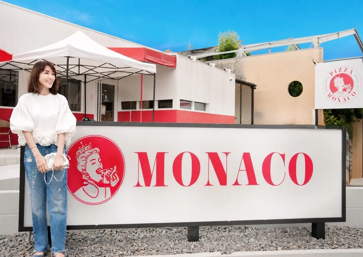 白い建物と「MONACO」の赤いロゴ看板の前。