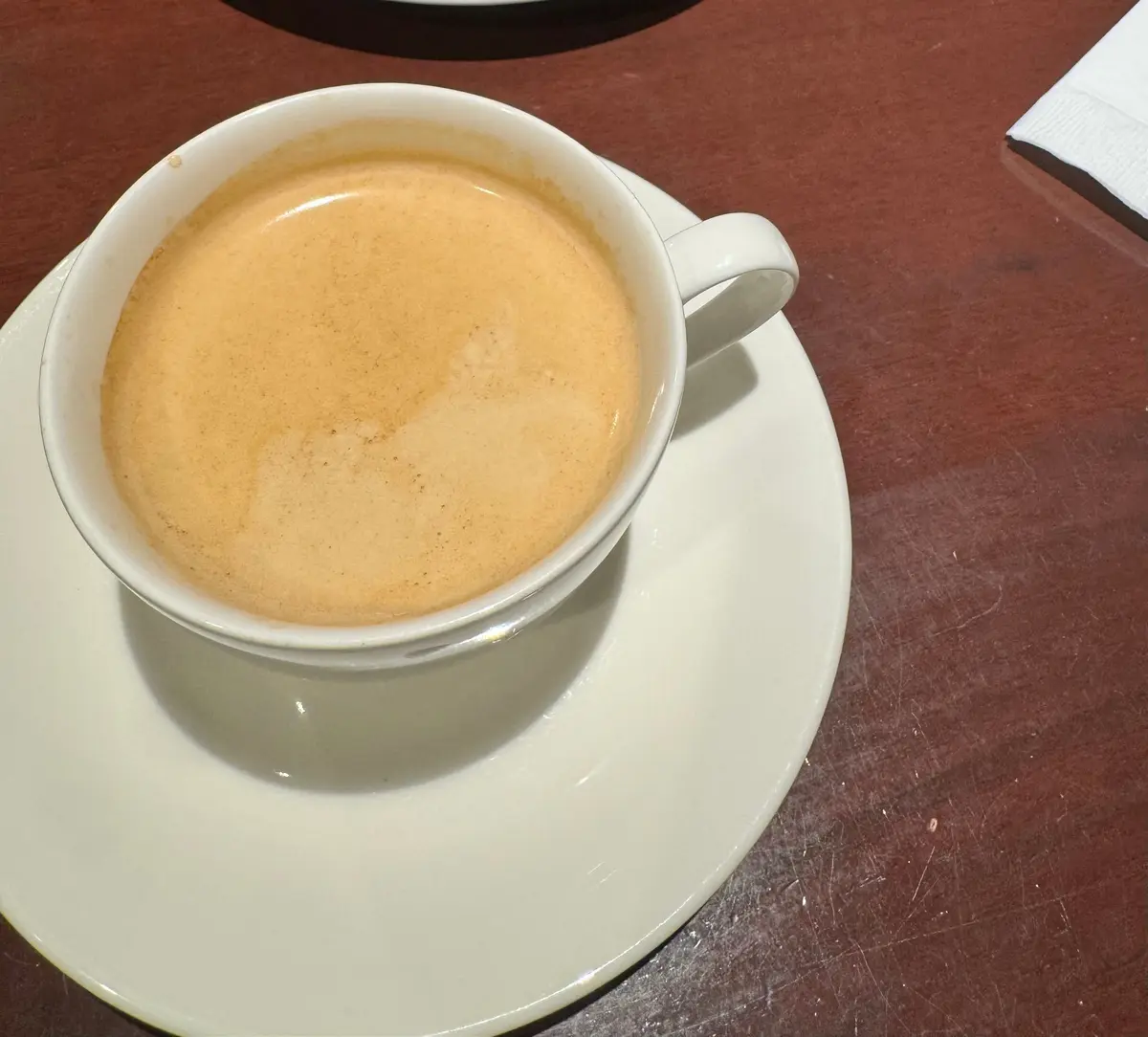 白い磁器のカップに入った、茶色のエスプレッソが置かれている。カップは白い皿の上にあり、木製のテーブルに置かれている。