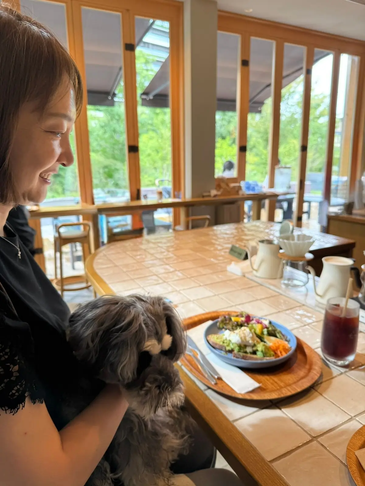 カフェのテーブルで、女性が灰色の犬を抱き、サラダと飲み物が置かれている。背景には大きな窓と緑が見える。