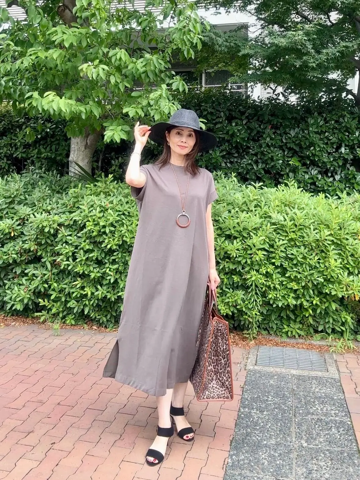 グレーのワンピースと大きな帽子を着た女性が、緑の低木に囲まれた通りに立っている。右手にはヒョウ柄のトートバッグを下げ、左手は頭に帽子をかぶせている。足元には黒のヒールサンダルを履いている。
