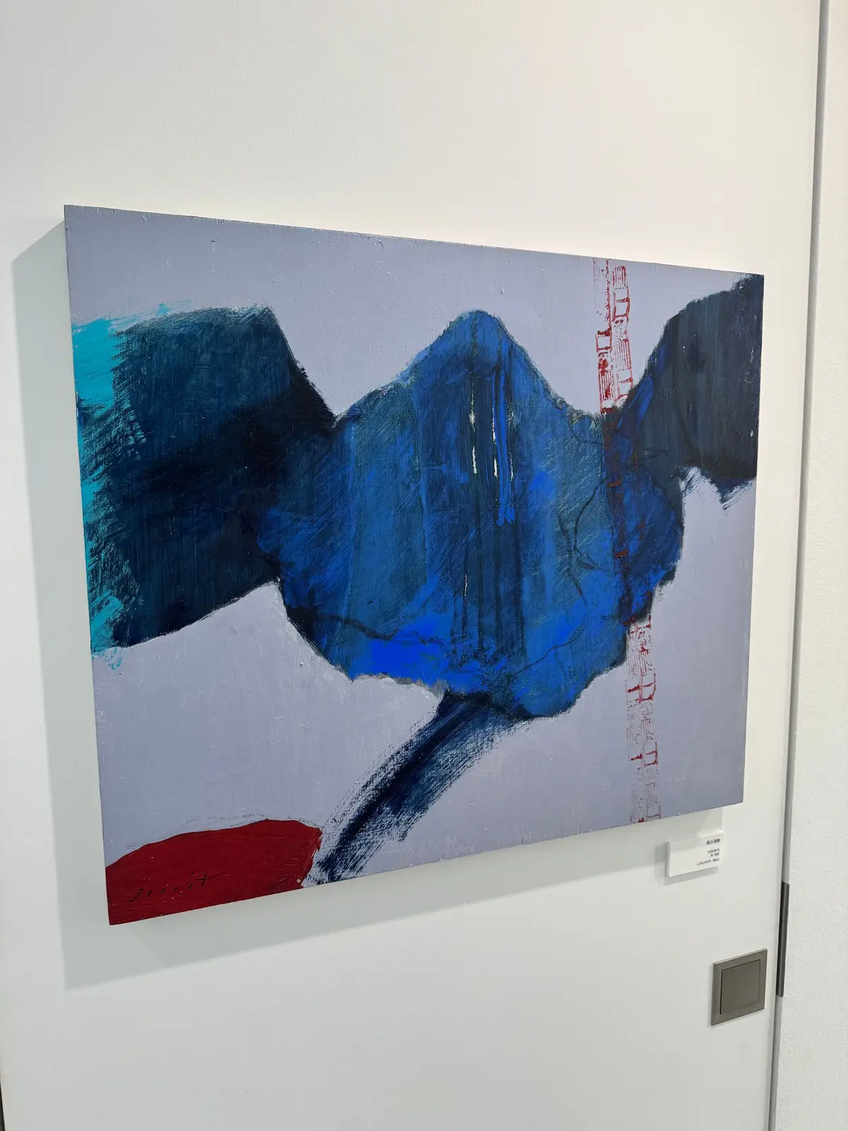 灰色の背景に、青と黒の抽象的な山のような形が描かれた絵画。下部には赤色の線が走り、右側には細い縦線が並んでいる。サインと思われる赤い文字が左下に小さく見える。