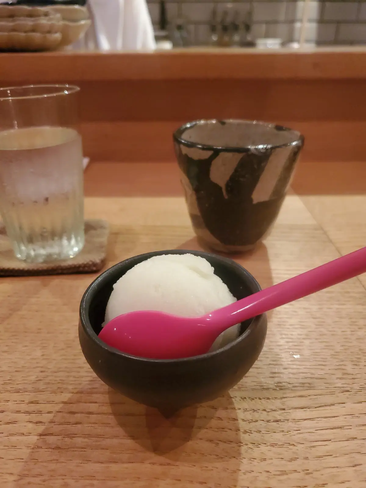 木製のテーブルに置かれた、黒い器に入った白いかき氷。ピンク色のスプーンが挿入されている。後ろにはグラスと茶碗が置かれている。