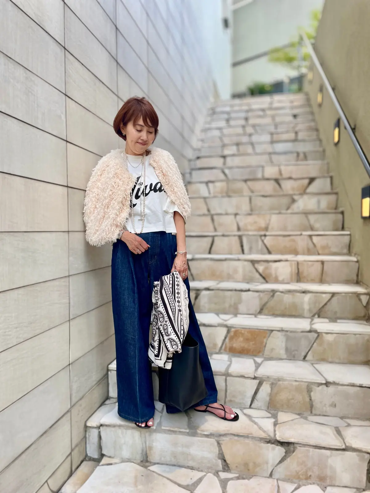 ベージュのモフモフのボレロ、白Tシャツ、藍色のワイドパンツを着用した女性が階段を降りている。黒いサンダル、パールネックレス、チェーンバッグ。背景は石積みの階段と壁。