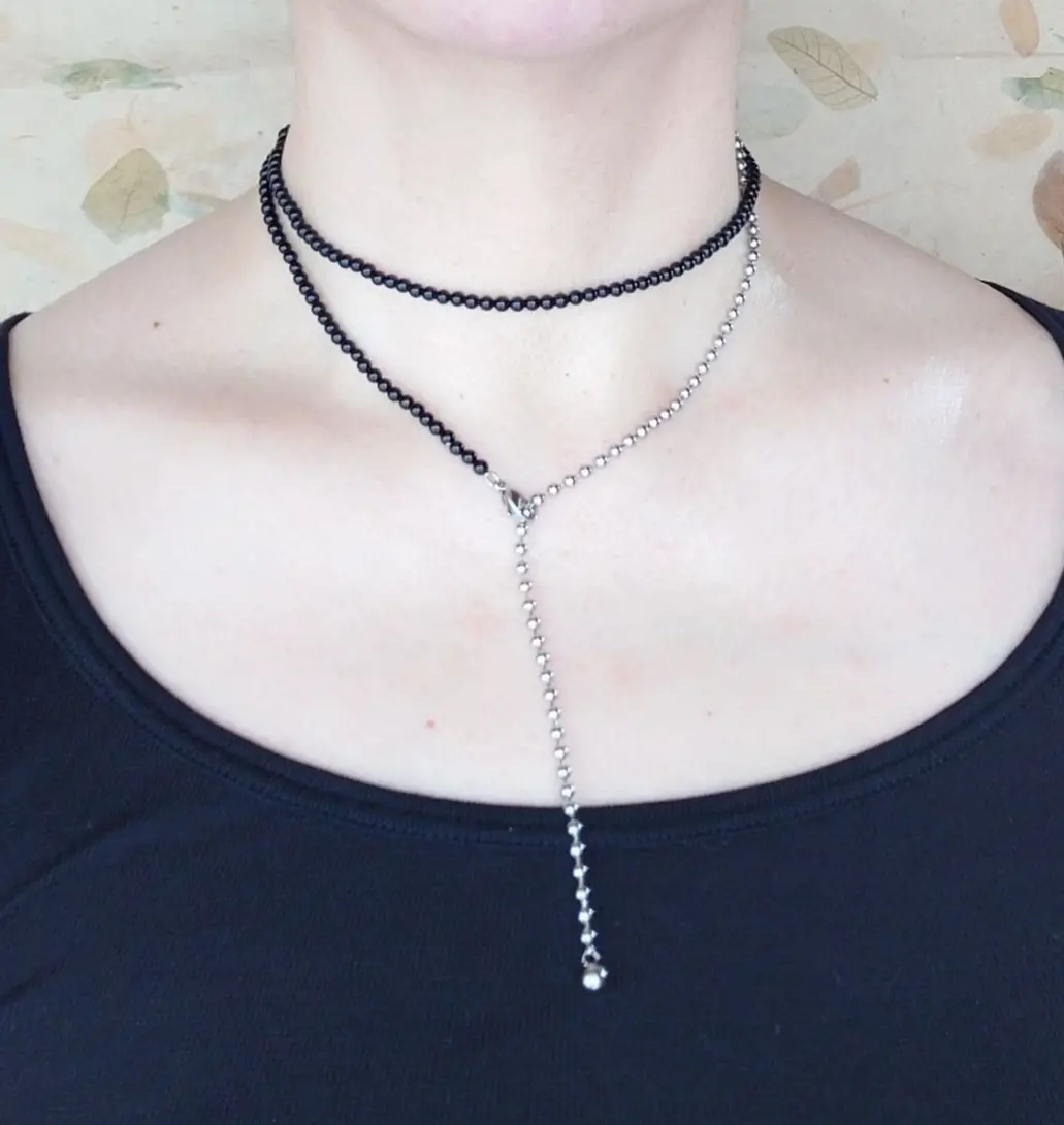 黒いカットソーを着た女性の首元。黒とシルバーの2連ネックレスを着用。上は細い黒いチェーン、下はビーズのチェーンが胸元でV字に重なっている。背景は葉の模様が描かれた淡い壁。