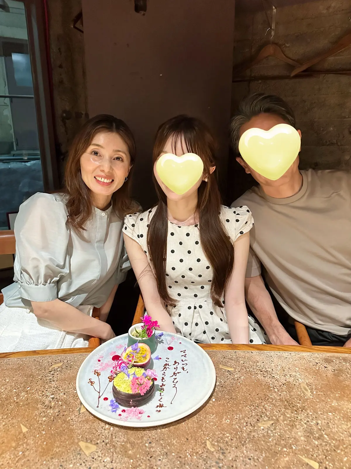 笑顔の女性3人が、花とフルーツを飾ったケーキを囲んでいる。左は白いシャツ、中央は水玉模様のワンピース、右は白のTシャツを着ている。背景には窓とレンガ壁が見える。