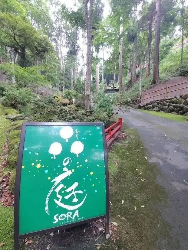 緑色の案内板と、その奥に続く木々の並木道。案内板には、白地に黒の筆文字と「SORA」のロゴが描かれている。背後には赤い手すりのある階段状の通路が見える。