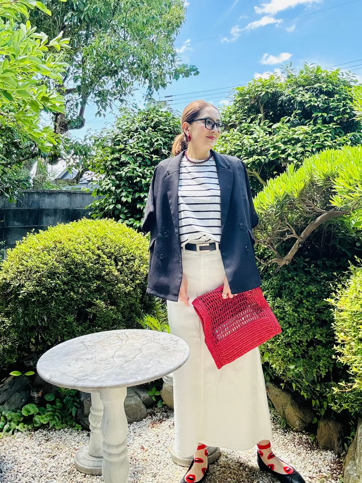 女性が庭でポーズし、黒のブレザージャケット、ストライプのトップス、ベージュのスカートを着て、赤の編みかごバッグを手にしています。背景には緑の低木と丸い石のテーブルが配置されています。