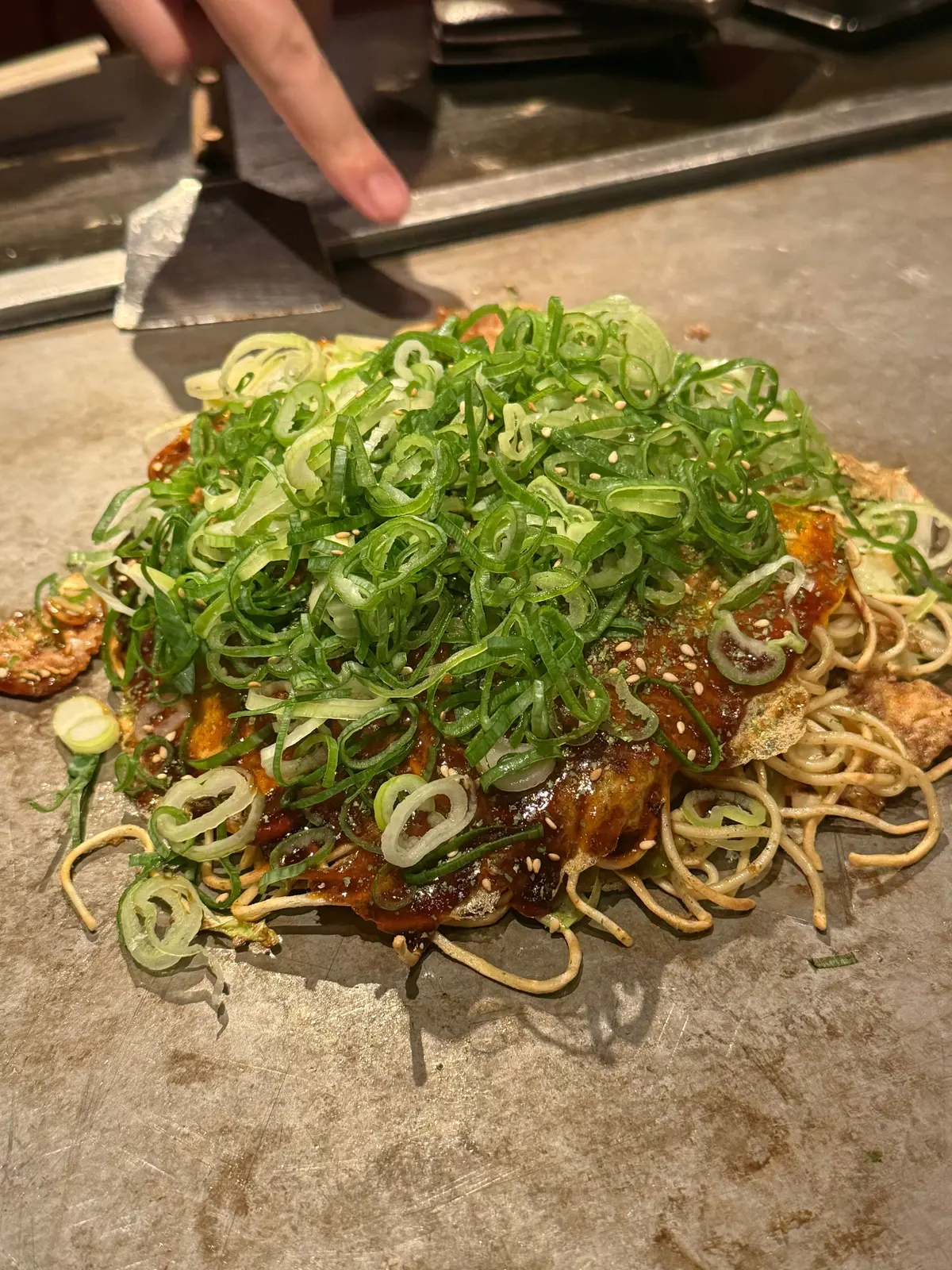 鉄板の上に置かれた、緑色のネギがたっぷりかかったお好み焼き。麺類や紅しょうが、甘辛いソースも見える。