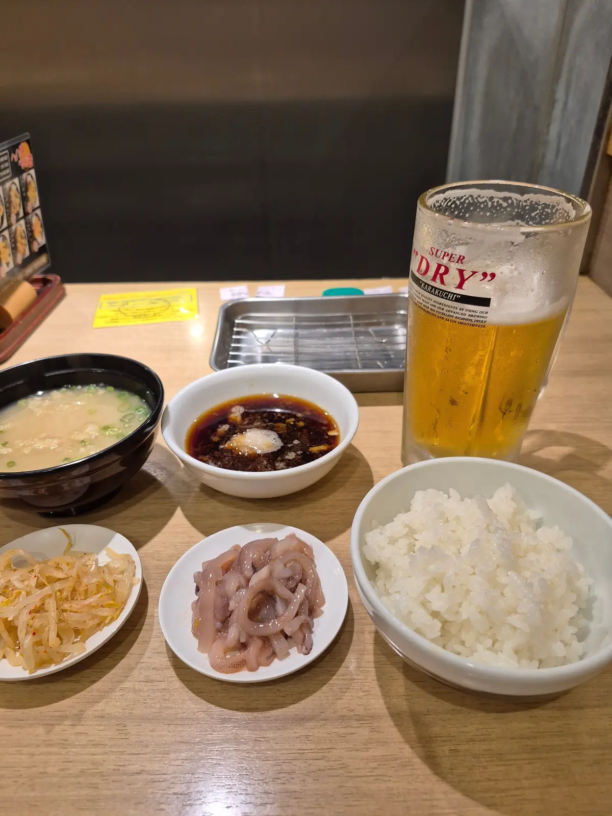 テーブルに置かれた日本の食卓。左上にはスープ、中央には焼肉の鉄板、右にはビール。その他、白米、ネギ、ホルモン、タレなどが盛り付けられた小皿が並んでいる。