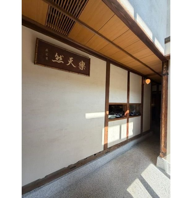 木造の庇（ひさし）の下に設置された、茶色の額縁に入った看板。「松天樂」と墨書された漢字が中央に配置されている。白い壁と木製の柱、窓枠が周囲を囲み、通路へと続いています。