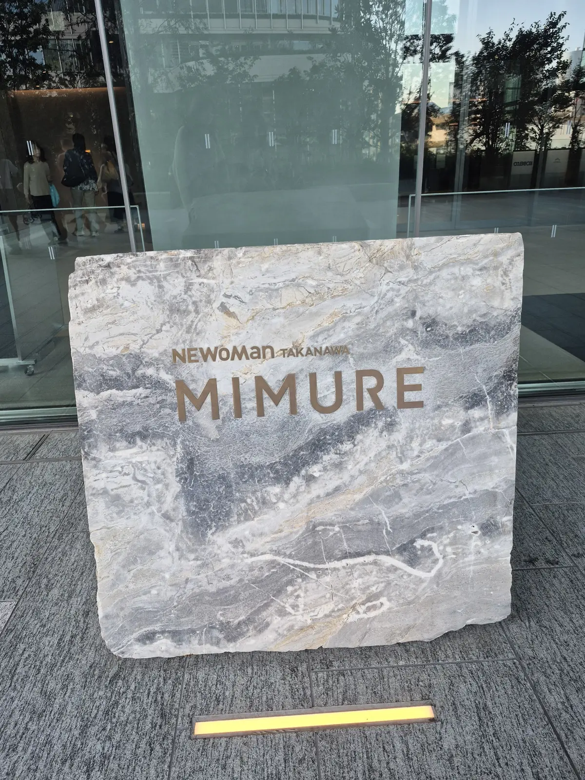 石材の看板に「NEWoMan Takanawa MIMURE」の文字が金色で刻まれている。背景にはガラス張りの建物と人々が見える。