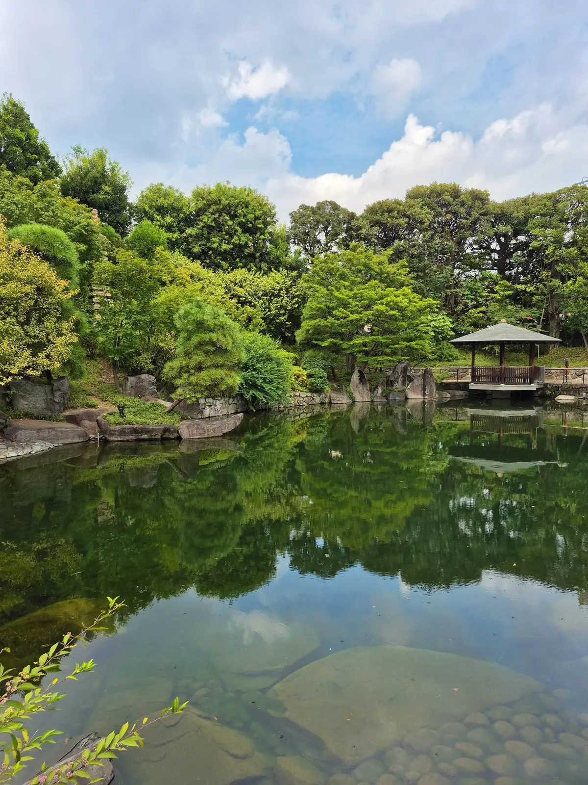 緑豊かな木々に囲まれた池。中央には大きな岩が配置され、その周りに水面が穏やかに波打っている。右端には木造の休憩所が見える。空は明るい青色で、白い雲が浮かんでいる。