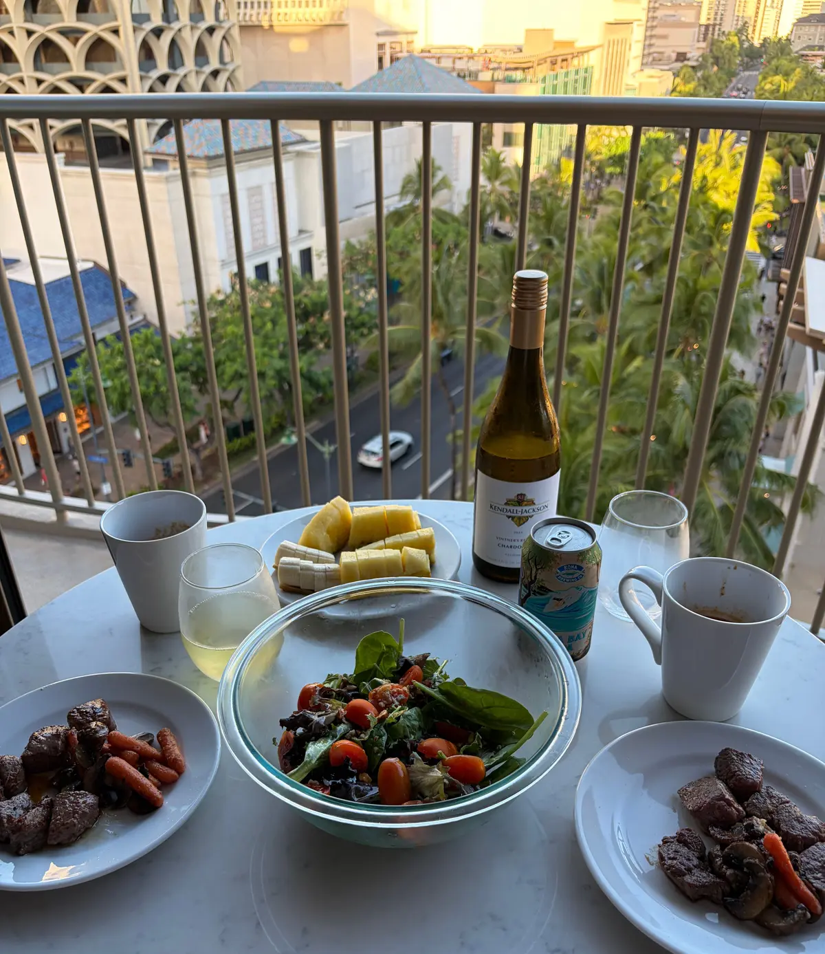 白いテーブルに、牛肉と人参、サラダ、チーズ、飲み物が置かれた食卓。背景には、夕暮れの街並みと高層ビルが見える。