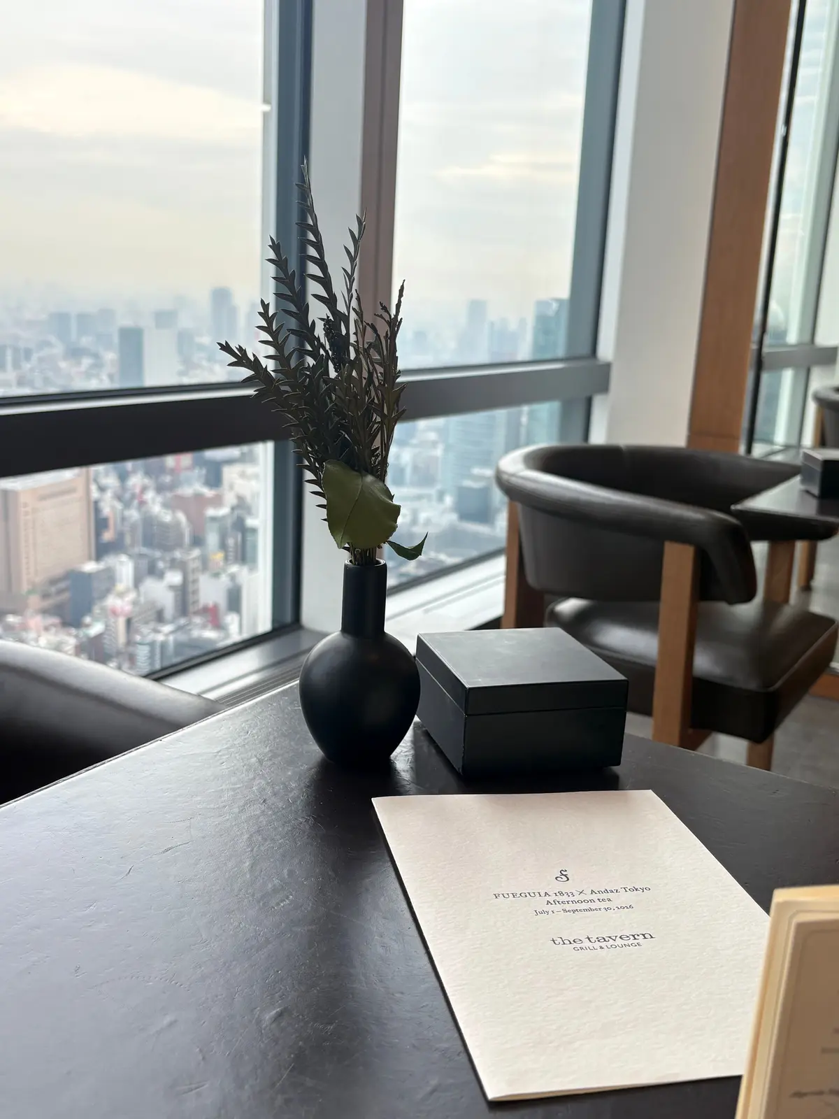高層階の窓から都市の景色が見えるテーブル。黒い花瓶に入った緑の植物、木製の箱、メニューが置かれている。