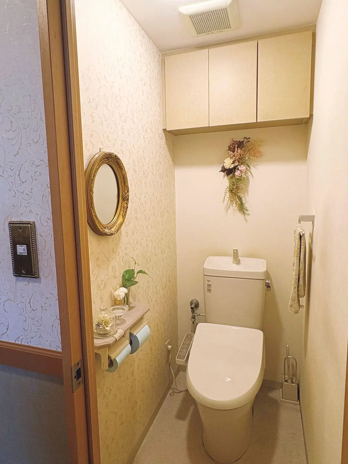 ベージュの壁紙を背景に、白い便器と上部収納が付いた洗面所。右壁には花束の装飾、左壁には鏡と観葉植物が置かれた棚がある。