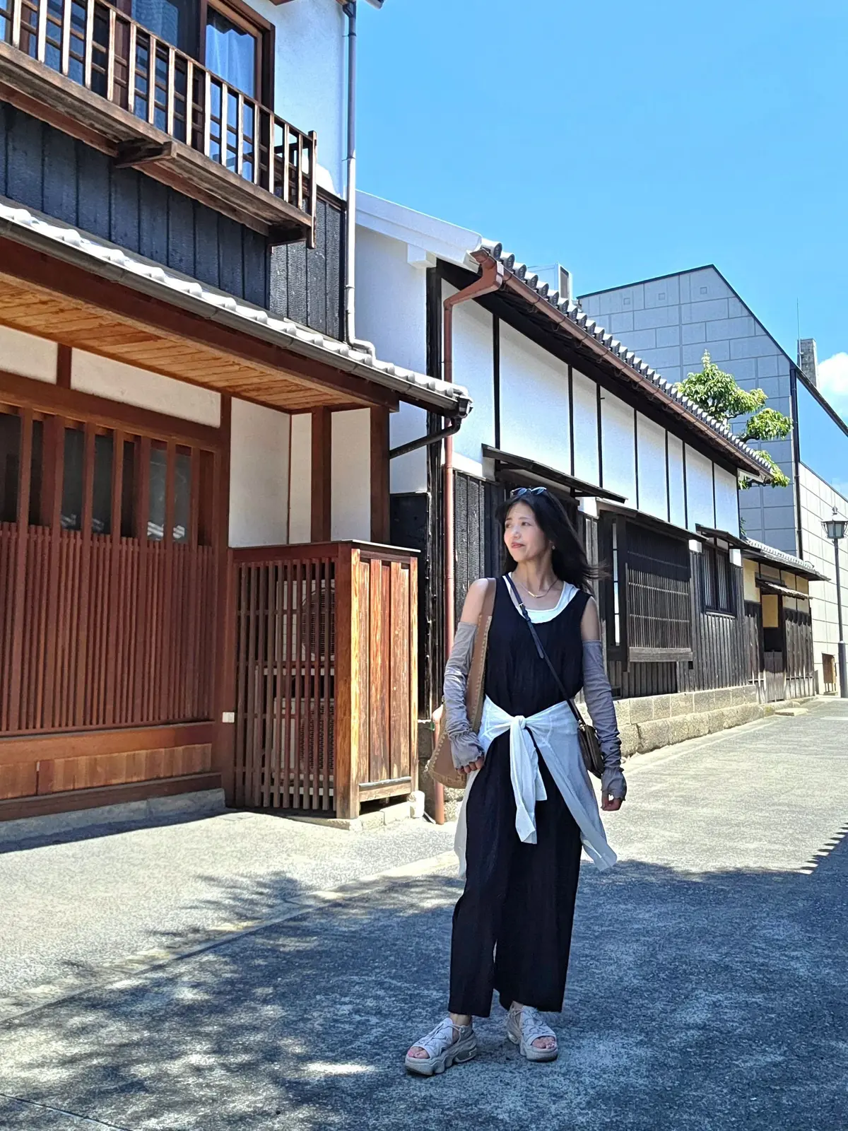 路地の奥に立つ女性。黒いタンクトップに白いリボンが巻かれたトップス、黒いパンツを着用。周囲には木造の伝統的な家並みが並び、青空が広がる。